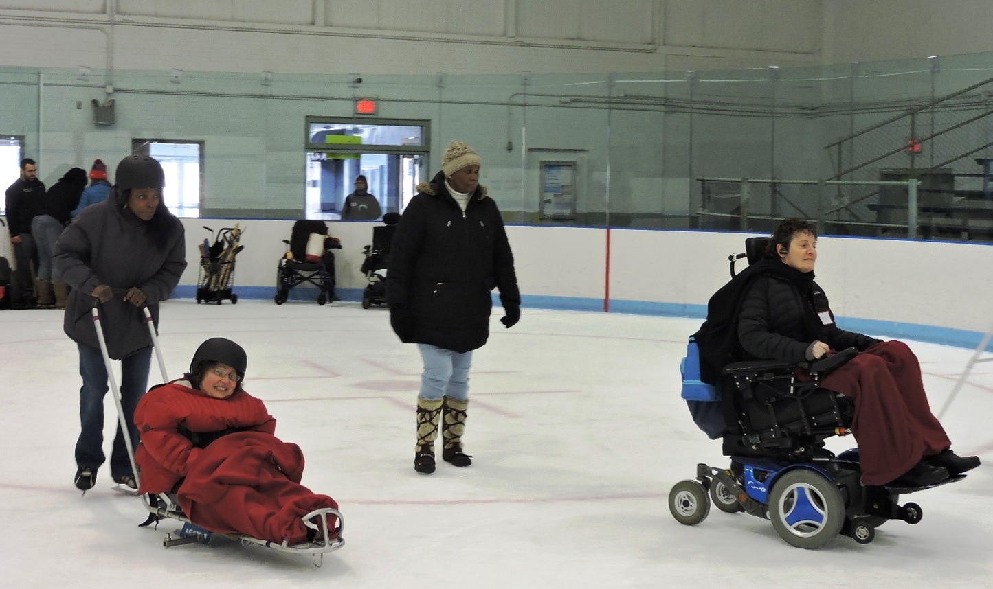 Two seated skaters and two standing skaters on the ice. The skaters are wearing jeans, winter coats, hats, and gloves. The seated skaters have blankets over their legs.
