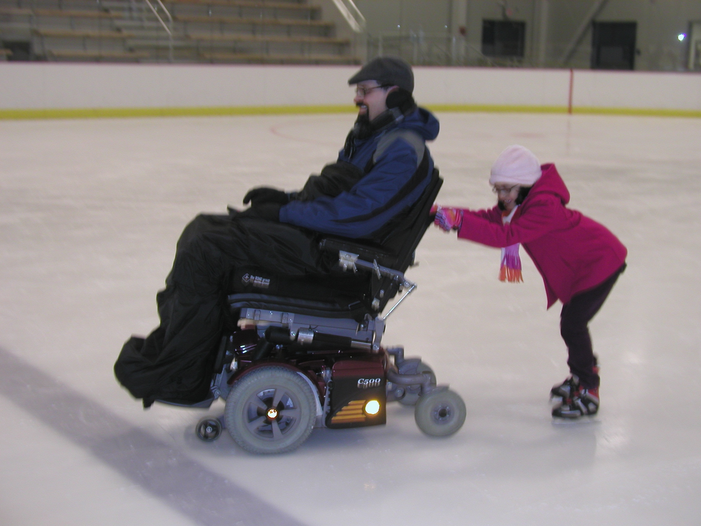 A power chair user is towing a child on skates, who is holding on to the back of the chair.