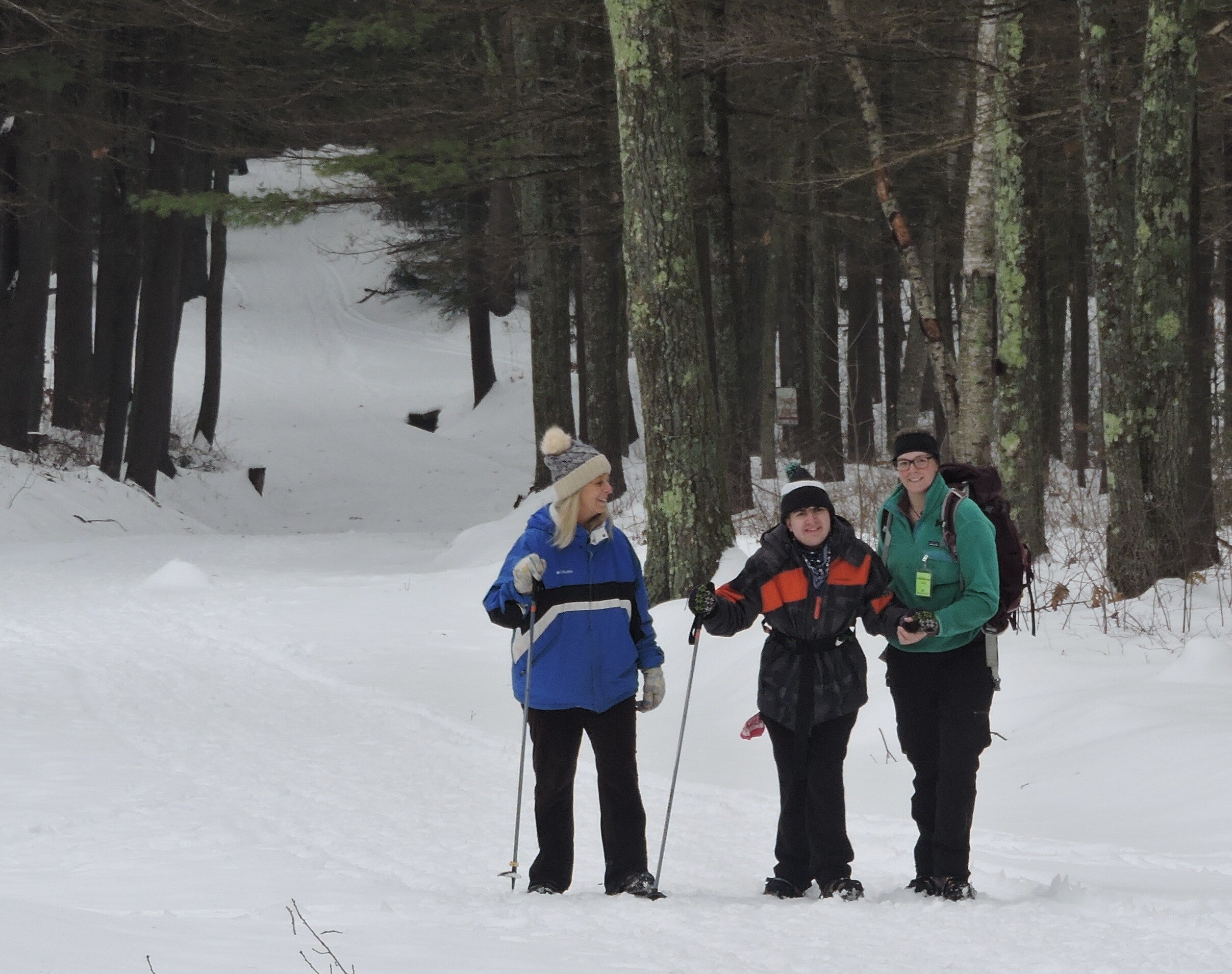A picture of three smiling people standing in the snow in winter clothes with trees behind them. Two are holding hiking poles. 