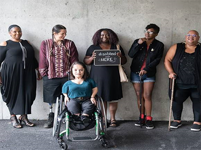 A group of people posing for a photo and holding a sign with the message, “disabled and here.” One person is sitting in a wheelchair.  