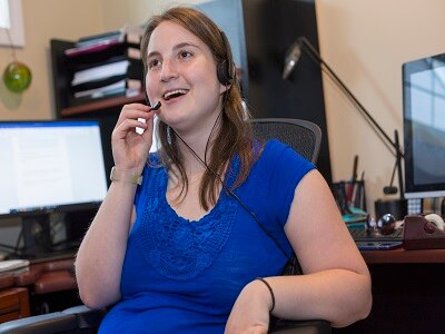A young white woman with cerebral palsy sits in an office with a headset on. She is turned away from her computer and has a happy look on her face.