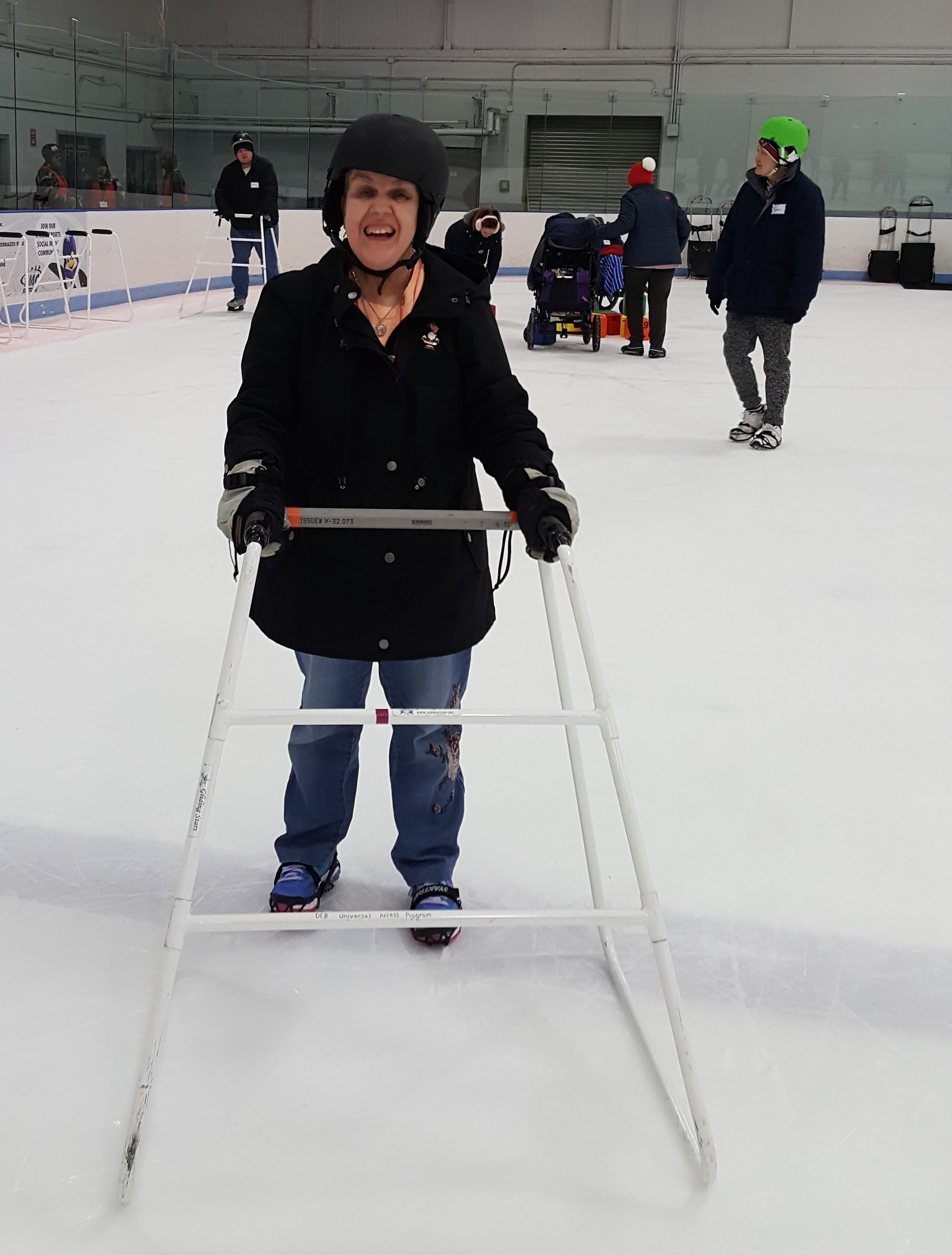 A person wearing a helmet and ice grippers is standing on the ice with a walker in front of them. A bar is across the back of the walker.