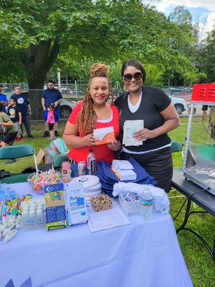 Two female staff members smile as they hold up COVID-19 test kit boxes near a table. 