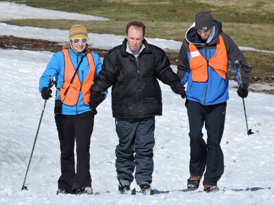 Two people wearing orange vests assist a skier.