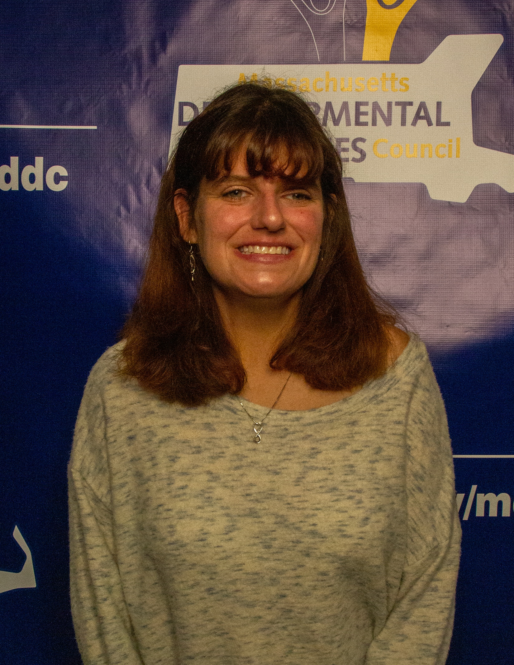 A white woman with medium length brown hair and bangs wearing a cream colored sweater stands in front of a blue banner with the MDDC logo on it.
