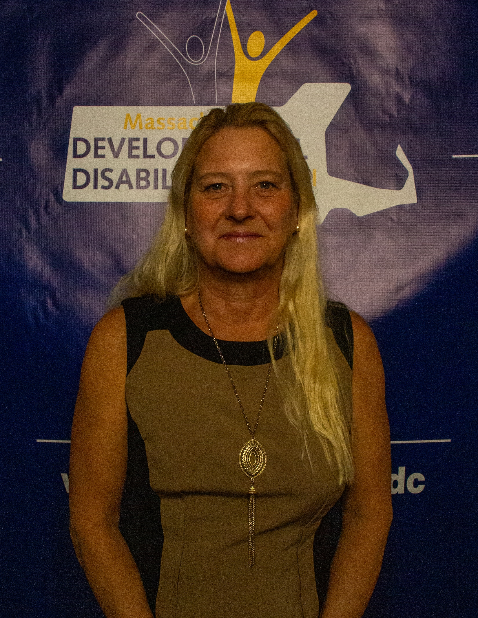 A white woman with long blonde hair wearing a beige and black dress stands in front of a blue banner with the MDDC logo printed on it.