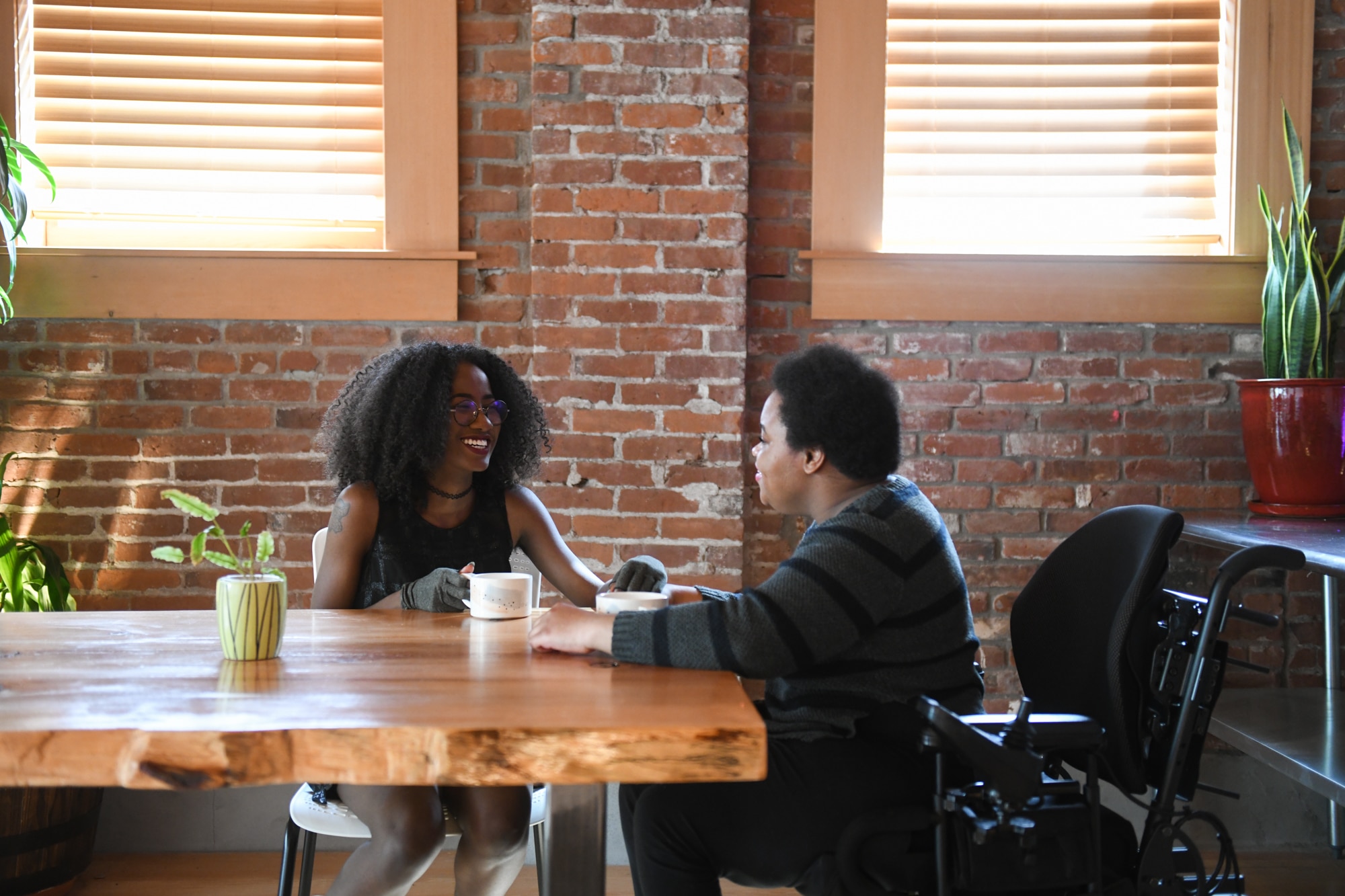 Two disabled Black people (a femme wearing compression gloves, and a non-binary person in a power wheelchair) smile and sit across each other while on a coffee date in a brick building.