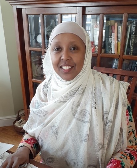 A black woman wearing a white headscarf and floral patterned shirt smiles while sitting in front of a bookcase.