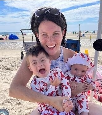 A white woman with dark hair wearing sunglasses on top of her head sits on the beach smiling with two small children in her arms.