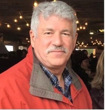 A white man with short gray hair and a mustache wearing a red and gray jacket smiling.