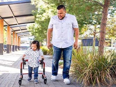 A toddler is walking with her assistive wheeled walker side-by-side with her male adult companion in a park.