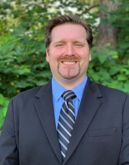 A white man with short brown hair and a beard wearing a blue suit and tie smiles in front of foliage.