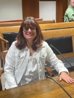 A white woman with long brown hair with bangs, wearing glasses and a white jacket sits in front of a microphone at a wooden desk.