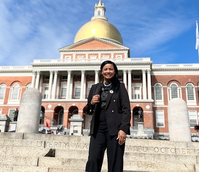 A latina woman with long black hair wearing a black suit stands smiling in front of the Massachusetts State House.
