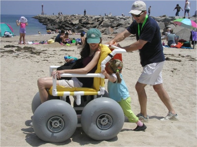 A man and a child push a woman in a beach wheelchair across the sand.