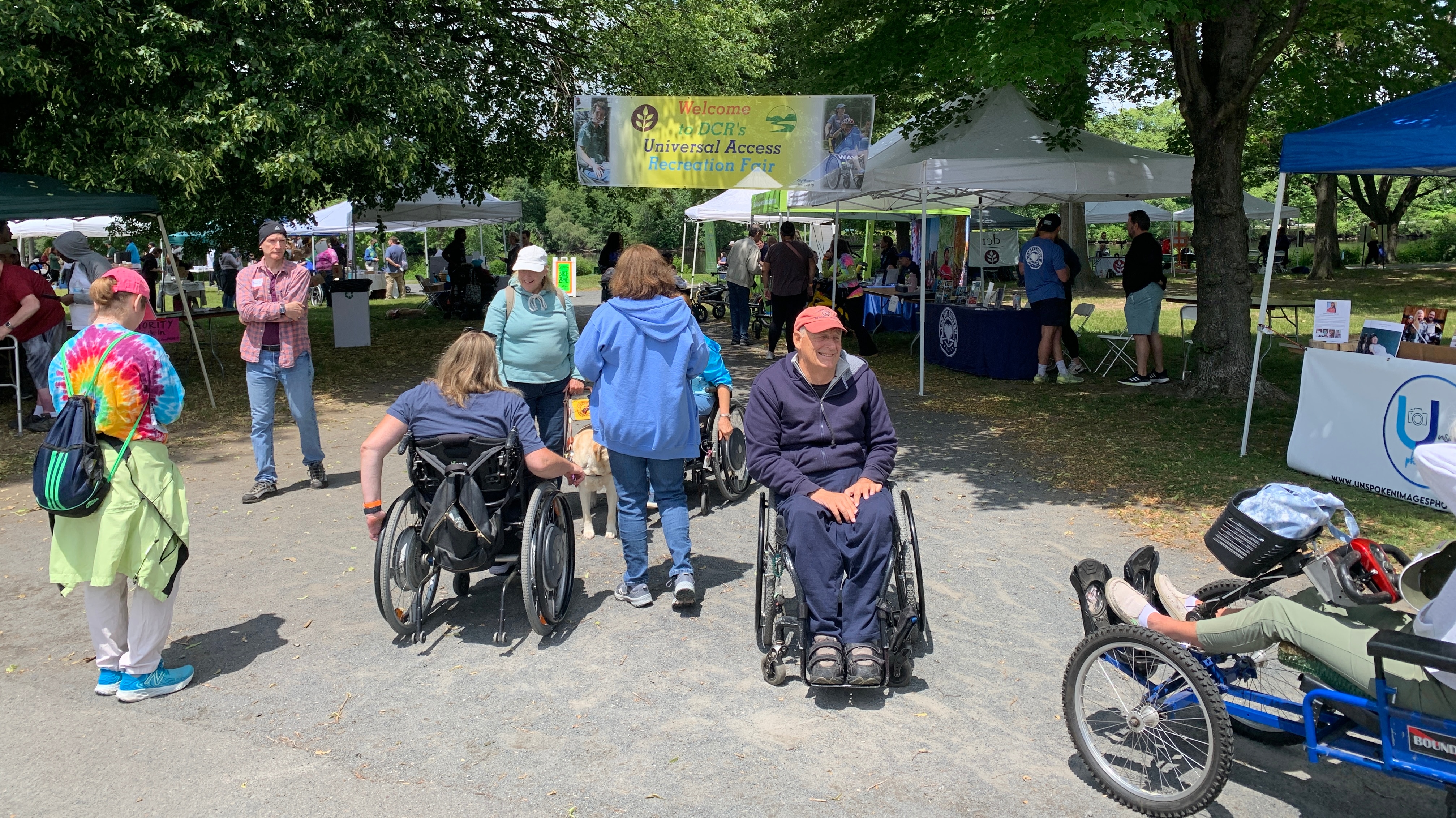 Tents are set up along a stone dust path that runs through a lawn surrounded by trees. There are information tables and equipment under the tents. Many people are are walking, using wheelchairs, and using adaptive cycles on the paths as they visit the tents.
