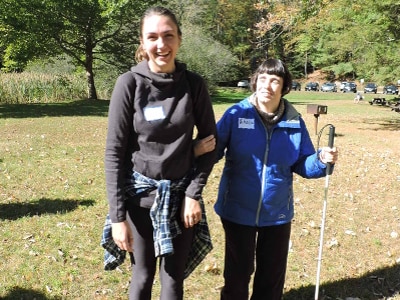 A hiker holding a white cane holds the arm of another hiker walking beside them.