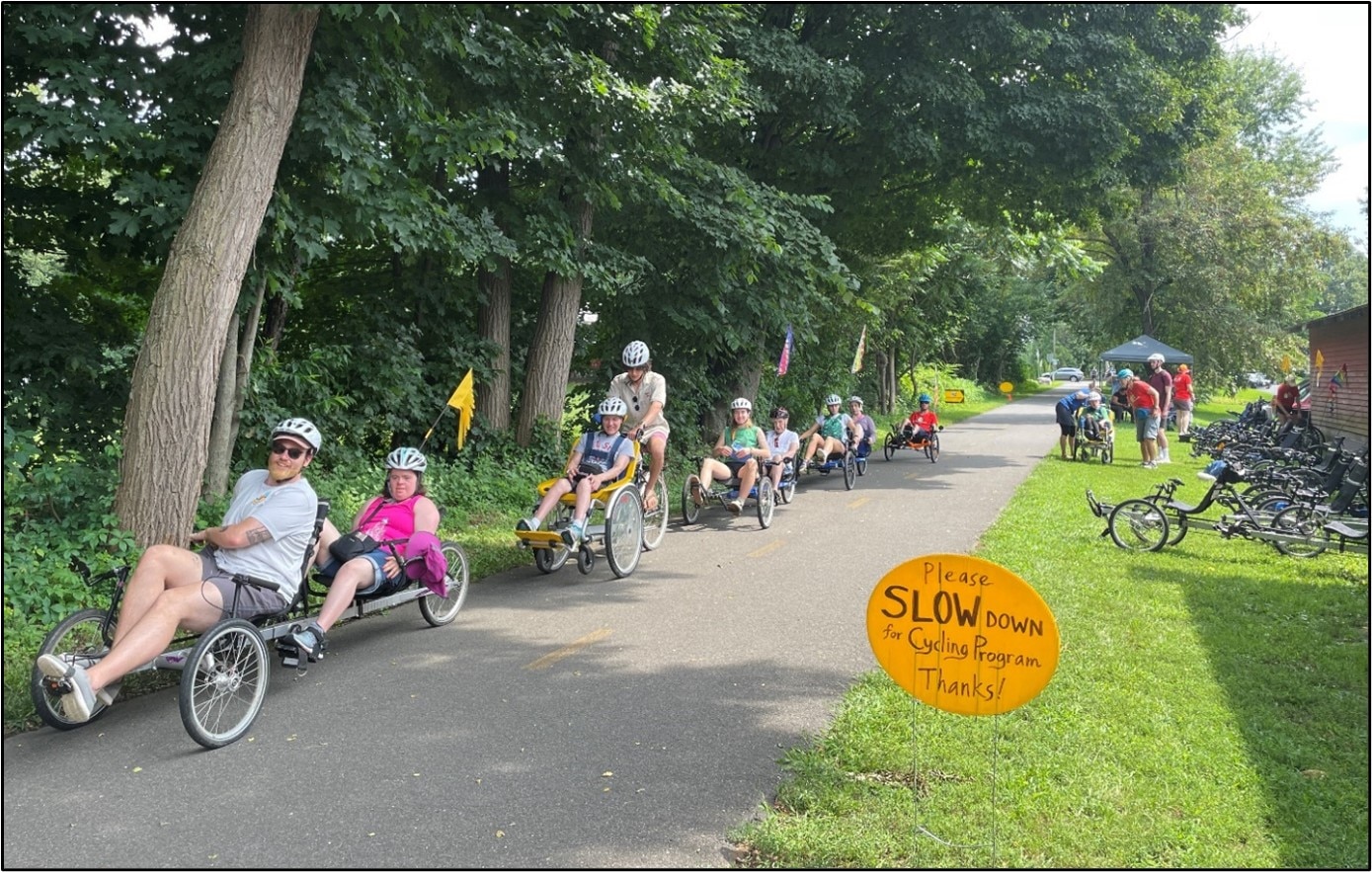 A group of bicyclists riding different types of bikes are travelling down a paved bike path.  There are a variety of bikes parked on the side of the path and a sign in front which says "Please Slow Down for Cycling Program. Thanks!"