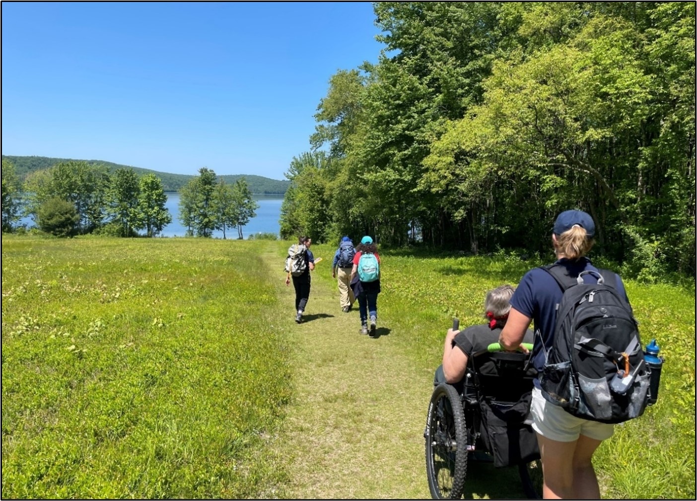 A group of hikers are walking towards a blue lake along a trail through a green meadow.  One person using a wheelchair is being pushed and 3 people are walking in front with back packs.  It's a sunny day and the meadow is surrounded by green trees.  
