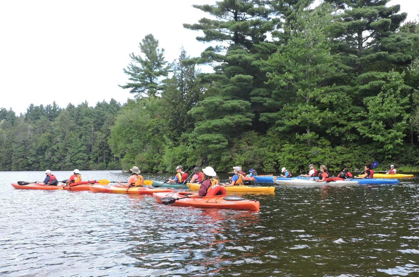 A photo of a large group of kayakers paddling close together. Some of the kayaks are single boats and some are tandem boats with two paddlers. Behind them is a wooded shoreline.
