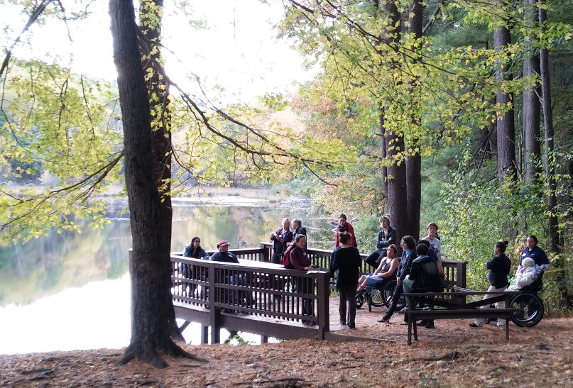 A photo of a viewing platform on the edge of a lake. A group of hikers is standing and sitting on the platform. Some of the hikers are using all-terrain hiking wheelchairs.