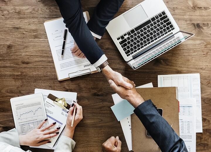 Overhead image of two people shaking hands over a table with a laptop and files of papers scattered about.