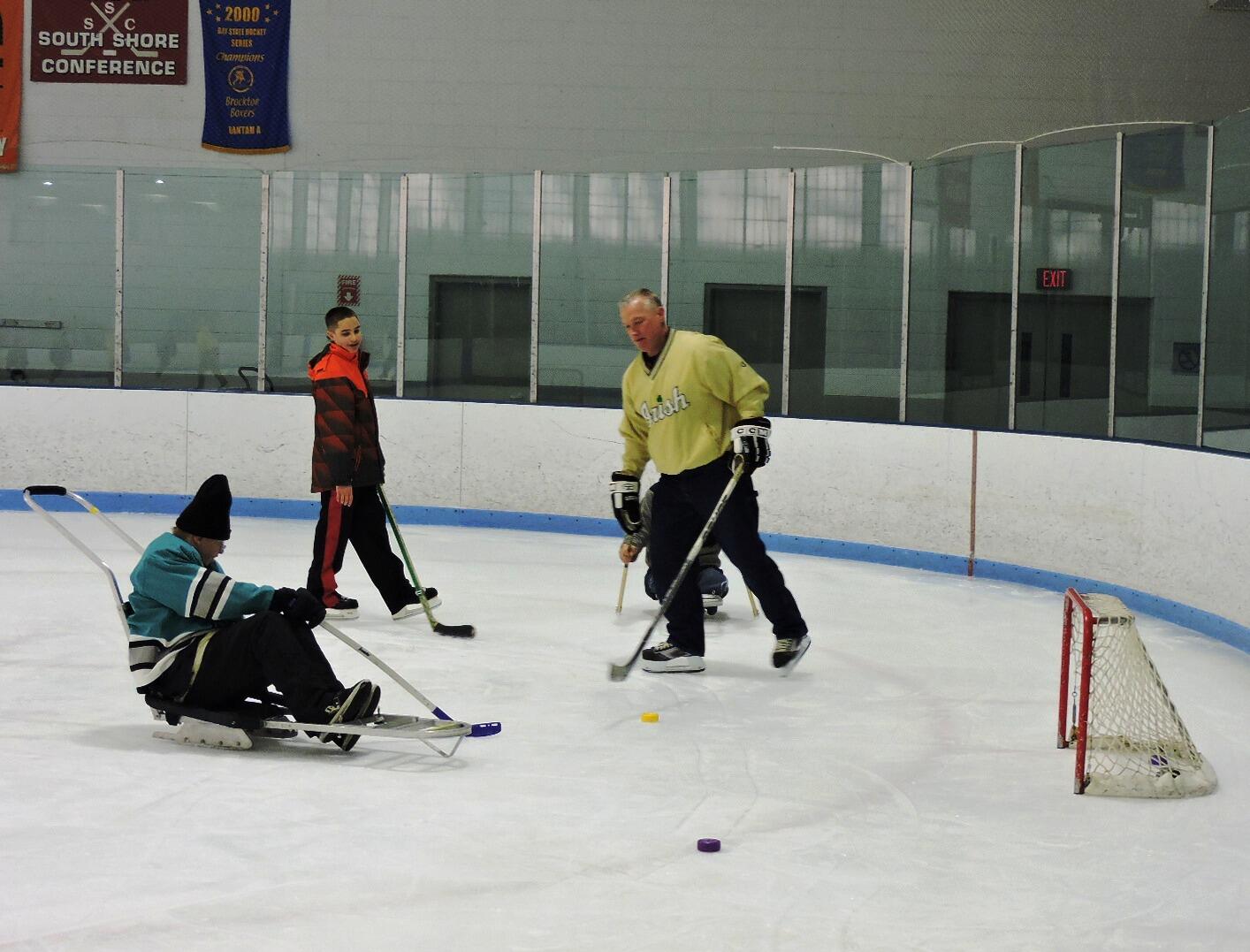 Four skaters are on the ice, facing a small hokey net. The skaters are shooting pucks towards the net. Two skaters are standing with skates on, and two skaters are sitting in ice sleds.