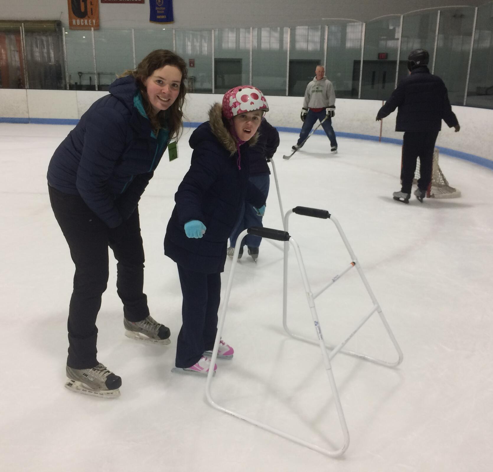 A young skater wearing a colorful helmet is standing in the foreground holding her arms out for balance. A skate walker is in front of her, and a staff member is standing behind her. They are both smiling at the camera. Behind them, two other skaters skate around a small hockey net.