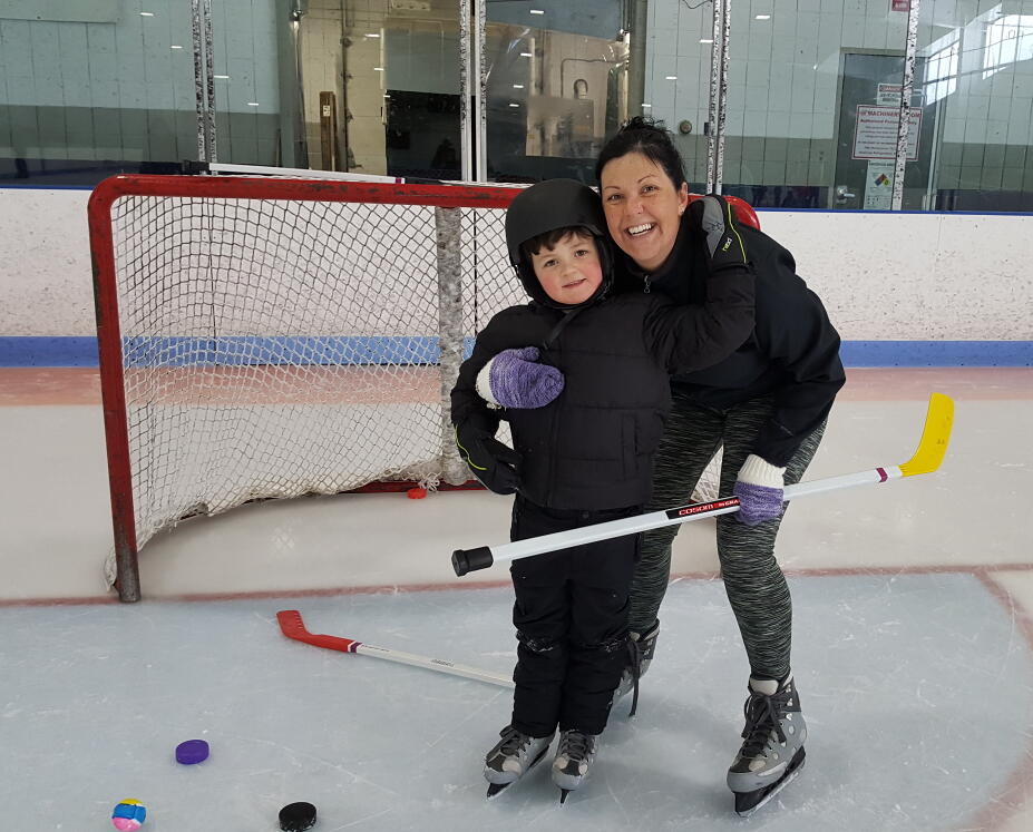 A woman and a child are wearing ice skates and standing in front of a hockey net. The woman is bending down and the two are hugging each other and smiling. There are colorful hockey sticks and pucks on the ice in front of them.