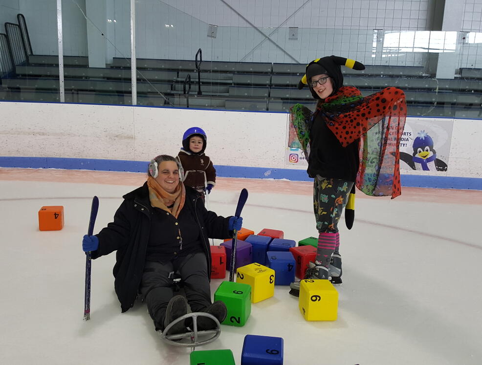 A woman in an ice sled, a girl on skates, and a young boy are standing in a pile of colorful blocks. They are smiling at the camera. The girl is spreading a colorful spotted cape with her arms, and wearing a hat with ears and a plush tail.
