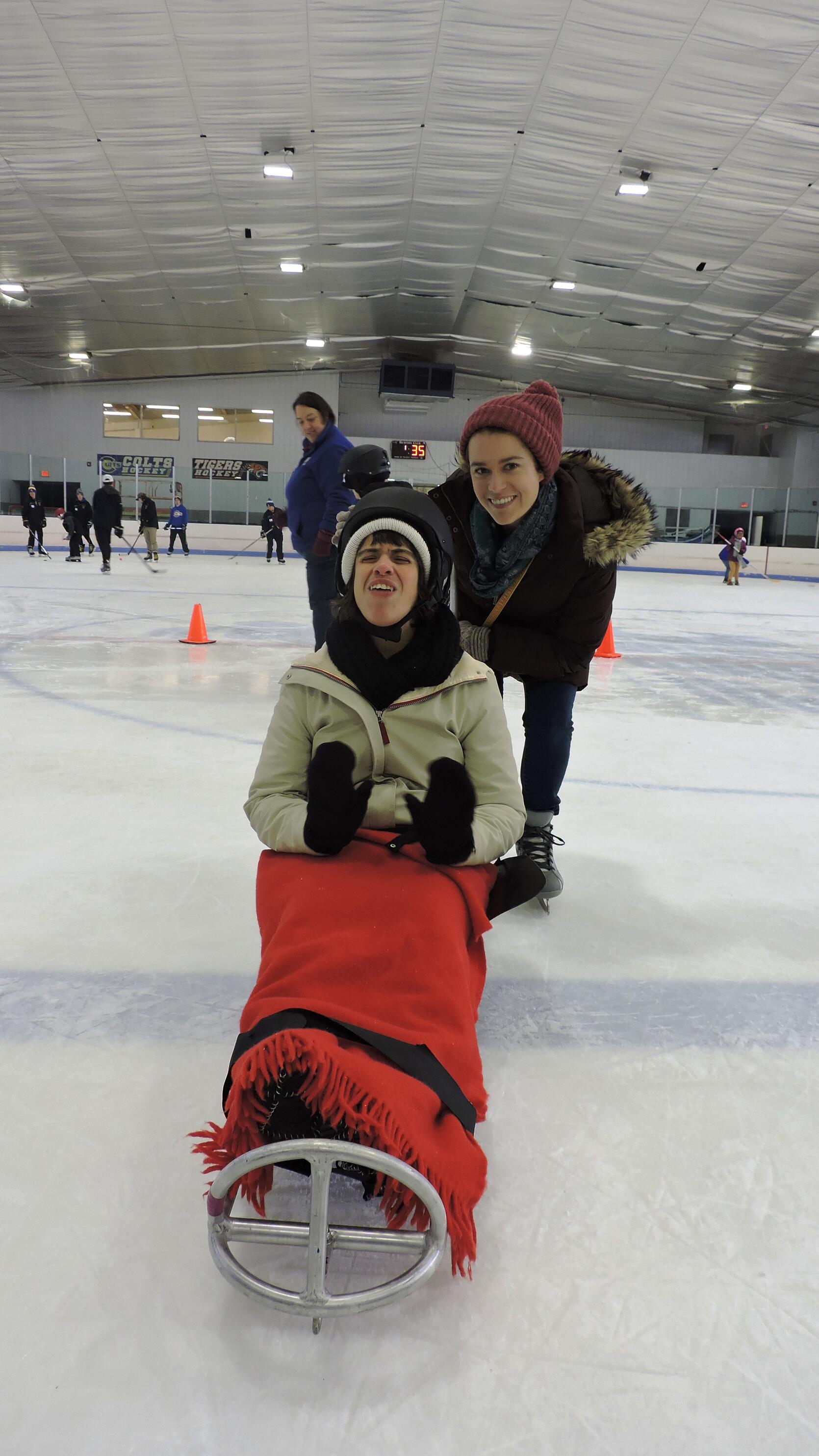 Two women are smiling at the camera, while a dozen skaters are visible on the ice behind them. One of the women is sitting in an ice sled with a blanket over her knees, thick mittens, and a helmet. The other woman is wearing skates and standing behind the sled, bending down.