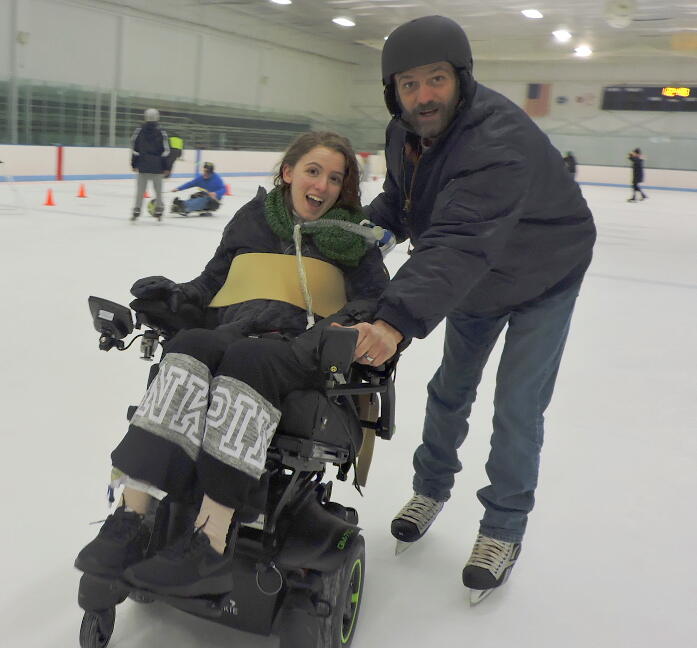 A woman and a man on the rink are smiling at the camera. The woman is in a power chair and the man is using ice skates and wearing a helmet. Behind them, a person in an ice sled is using a hockey stick to pass a large ball to a standing skater.