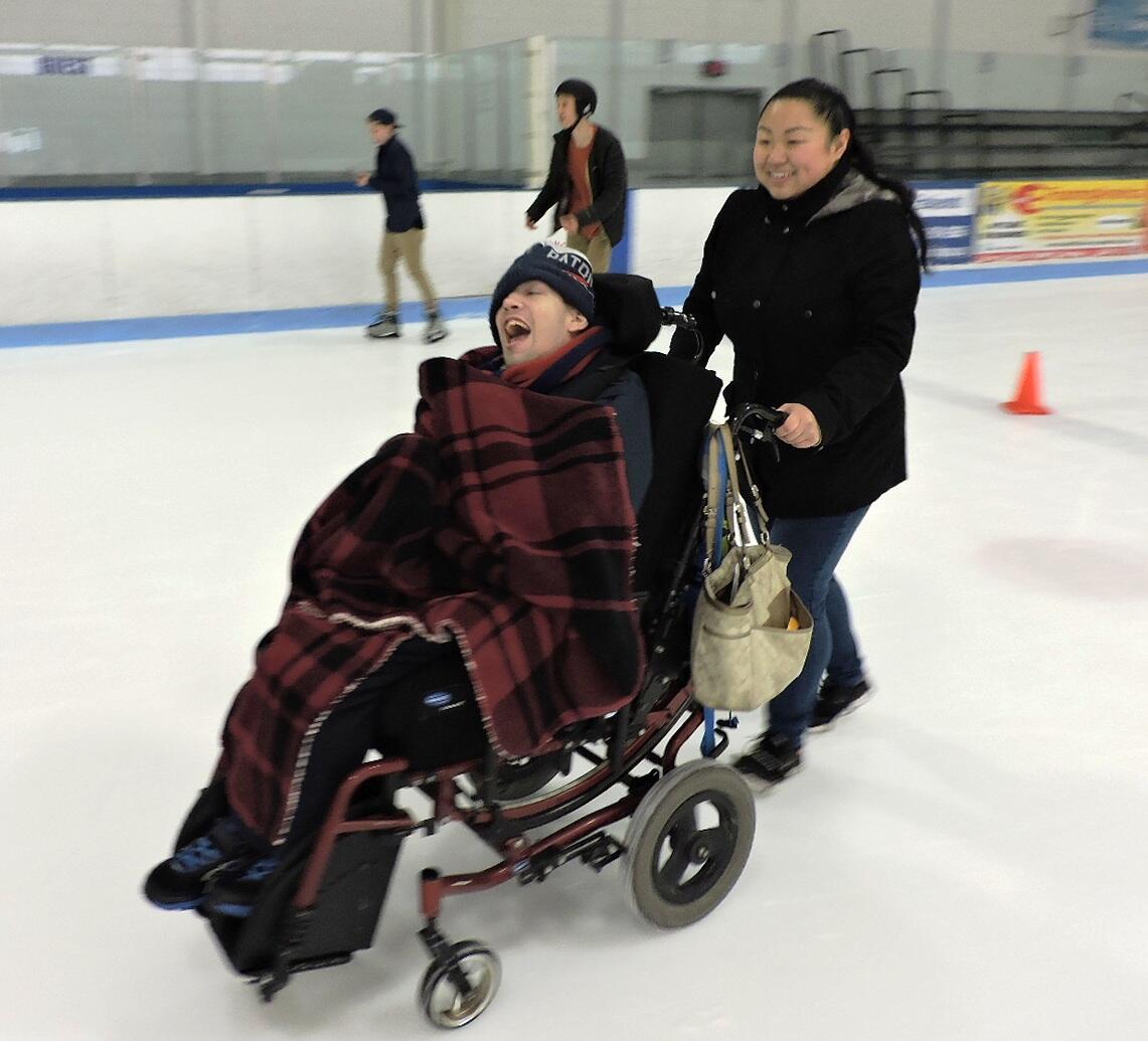 A person wearing ice grippers is pushing another person in a manual wheelchair across the ice. The person in the chair is covered in a plaid blanket and a warm hat. They are both laughing and smiling. Two standing skaters are skating by them in the background.