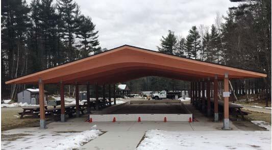 A pavilion with a wide roof sits in a grassy field dusted with snow. The pavilion has a concrete pad and path leading up to it. An ice rink is set up under the roof.
