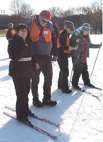 Two standing skiers are being guided across the snow by volunteers. One of the skiers is using ski poles, and one skier is not.