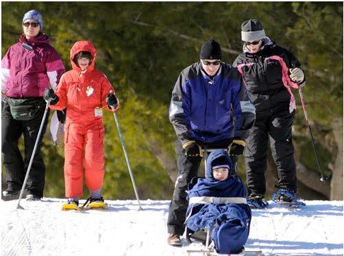 Two people are using snowshoes and ski poles on a snow-covered field. In front of them, a sit skier is wrapped up in a blanket, with a belt around their waist. The skier is being guided down a gentle slope by a person standing behind them and holding on to the sit ski.