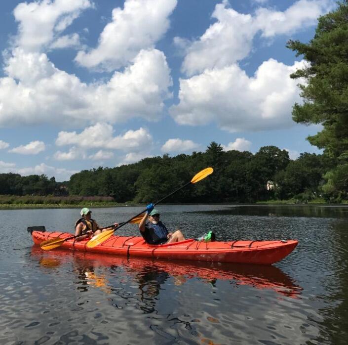 Two people are sitting in a tandem kayak with a closed cockpit. Both people have paddles. The person in front is raising their paddle into the air with one hand.