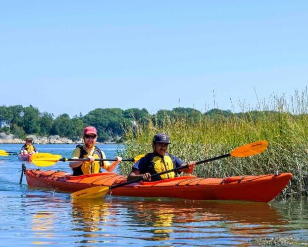 A photo of two people in a tandem kayak with a closed cockpit. Both people are paddling. Behind them is another tandem kayak with two paddlers.