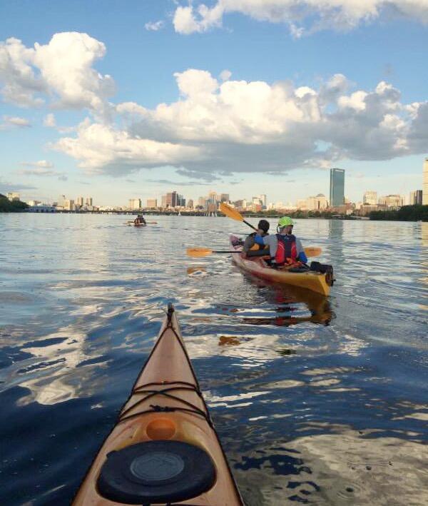 A photo taken from a kayak, showing two tandem kayaks ahead on the river. The kayaks are heading towards the Boston skyline.