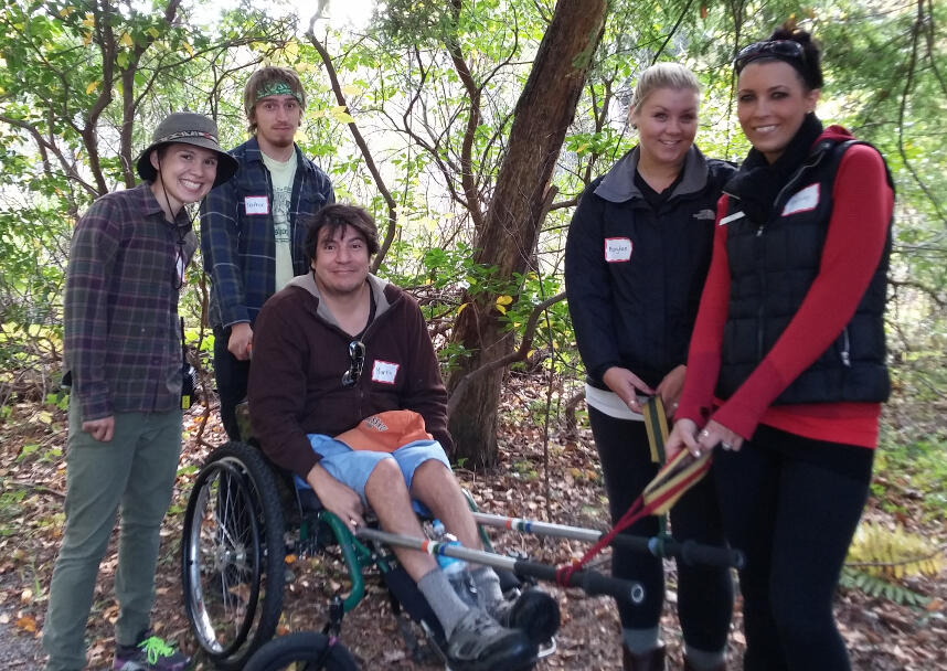 A group of hikers standing in the woods. One of the hikers is using an all-terrain hiking wheelchair with rickshaw poles on the front.