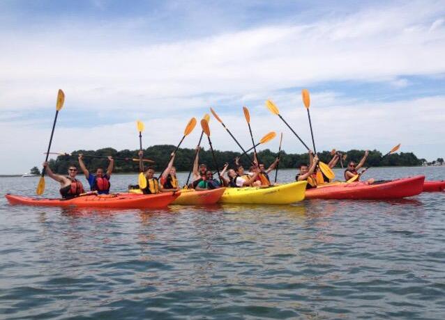 A group of thirteen kayakers are on the water. The kayakers are grouped close together and are raising their paddles straight up in the air and smiling.