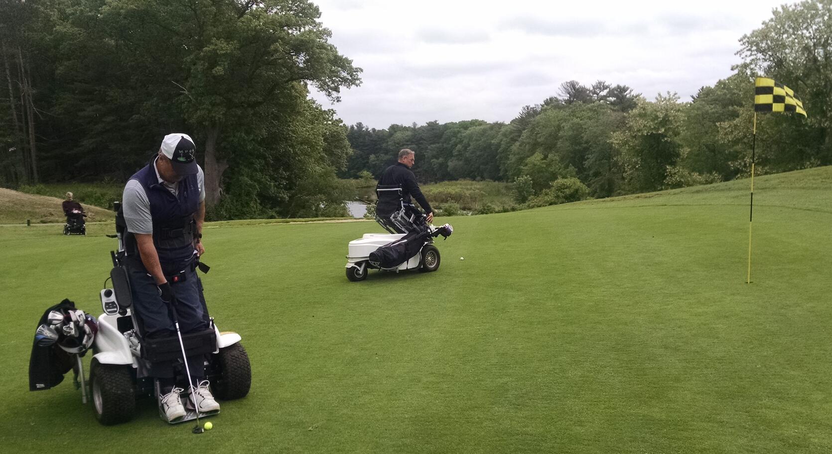 Two men are riding Paragolfers on a golf green near the hole. The men are raised up in the Paragolfers and setting up swings. Another man in a conventional power wheelchair is behind them. Water is visible in the distance.