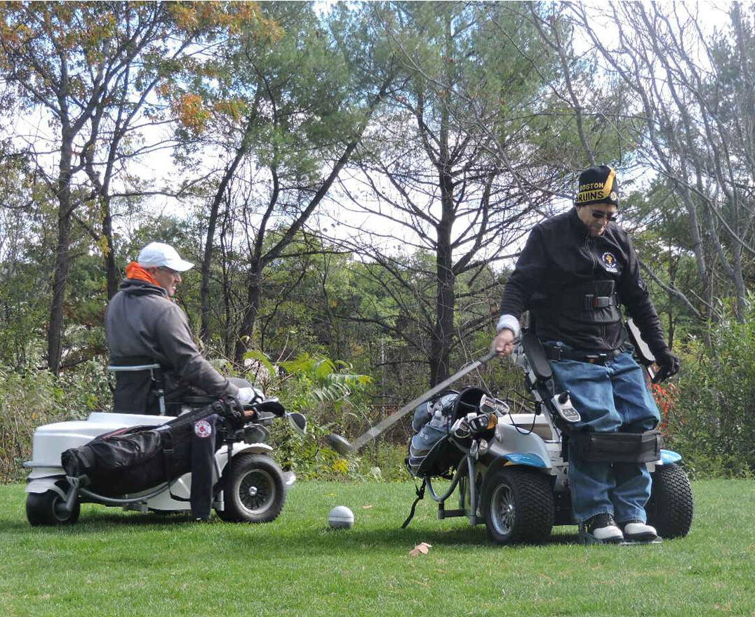 Two men are using Paragolfers on the grass. One man is raised up and swinging his golf club. A 5-inch diameter ball is on the grass. There are trees behind them.
