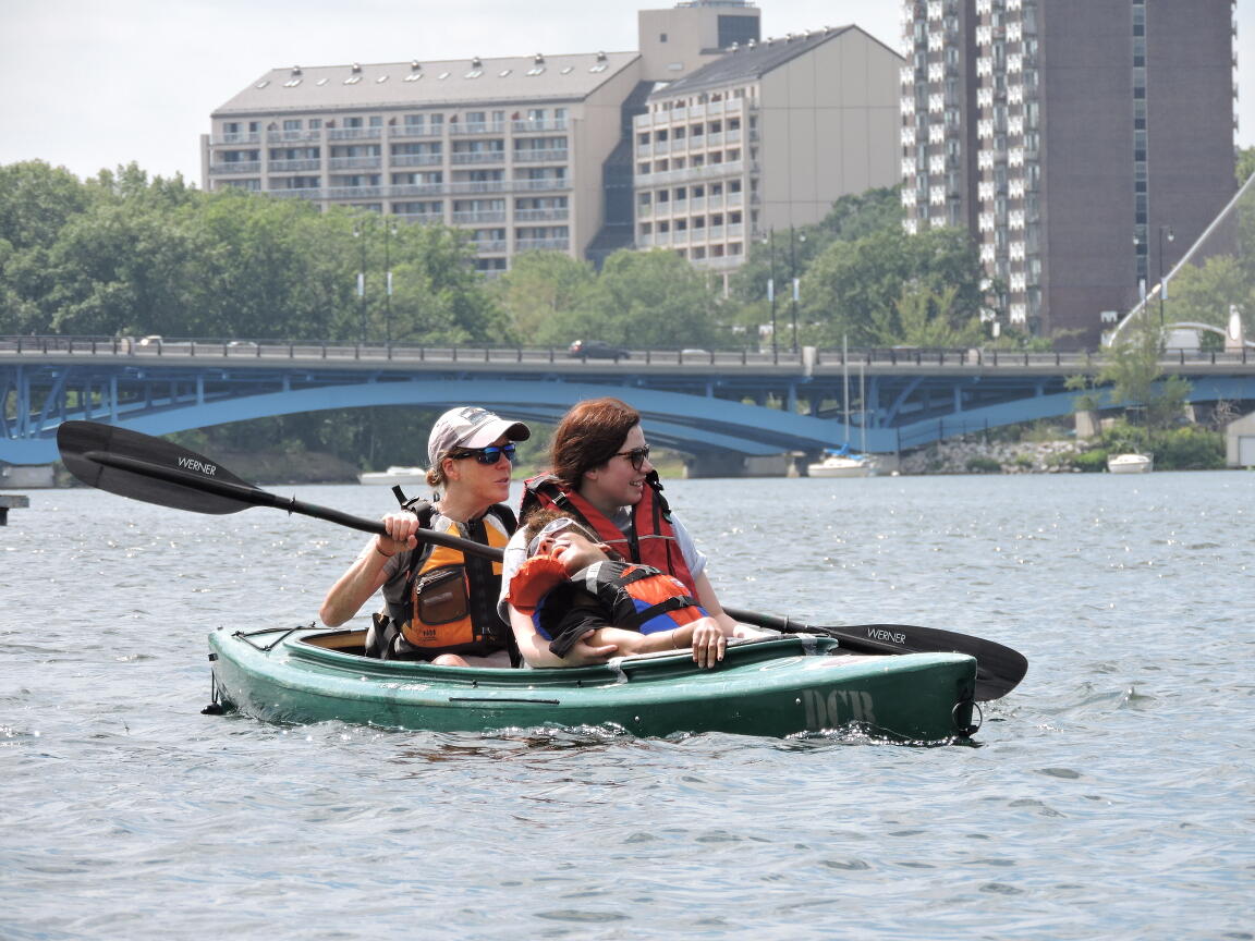 Three people are paddling in a tandem kayak. One person is sitting on the lap of another person in the front of the boat. The person in back is paddling. Behind them, The Burns Memorial Bridge and some high-rise buildings are visible.