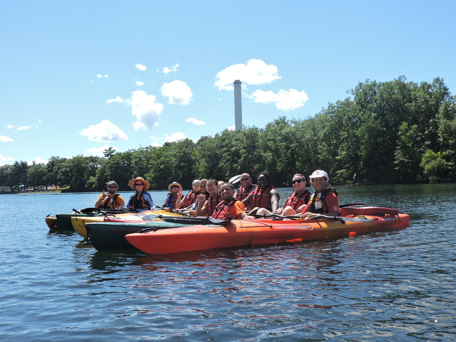 A dozen kayakers are grouped together on the water. Some of the kayakers are in tandem boats and some are in single boats. On the shore behind them, a large chimney is rising through the trees.