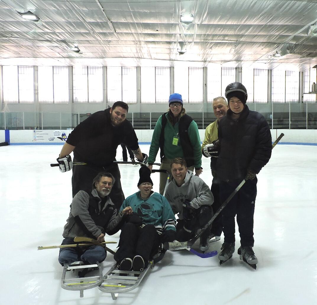 A group of skaters on the ice is smiling for the camera. Some skaters are using sleds, and some skaters are standing. Several skaters are holding hockey sticks.