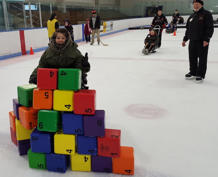 A woman wearing a thick faux fur hood is crashing through a tower of colorful blocks in her power wheelchair.