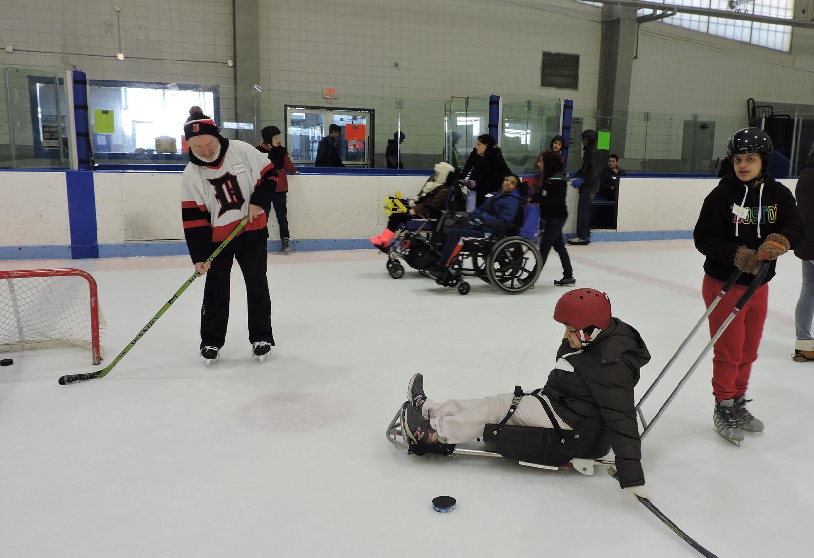 A skater in an ice sled is setting up to shoot a hockey puck at a small hockey net. Another skater is holding onto the handle of the ice sled. The goalie at the net has a stick out to catch the puck. In the background, two skaters in manual wheelchairs are being pushed across the ice.