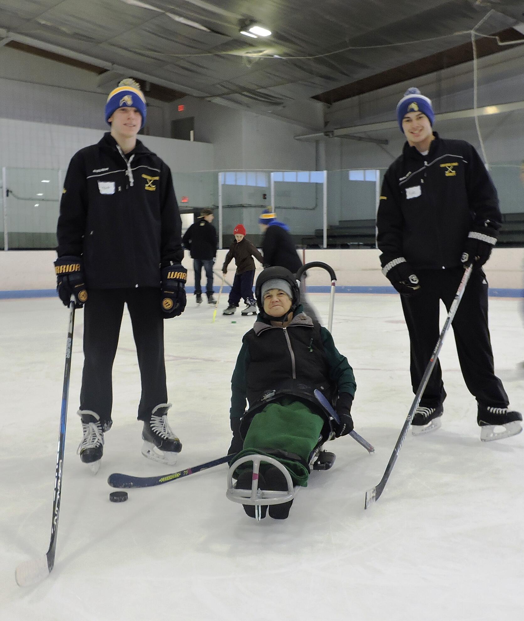 Two Chicopee Colts hockey players are standing on either side of a woman using an ice sled. The woman has her hockey stick lined up to shoot a puck.