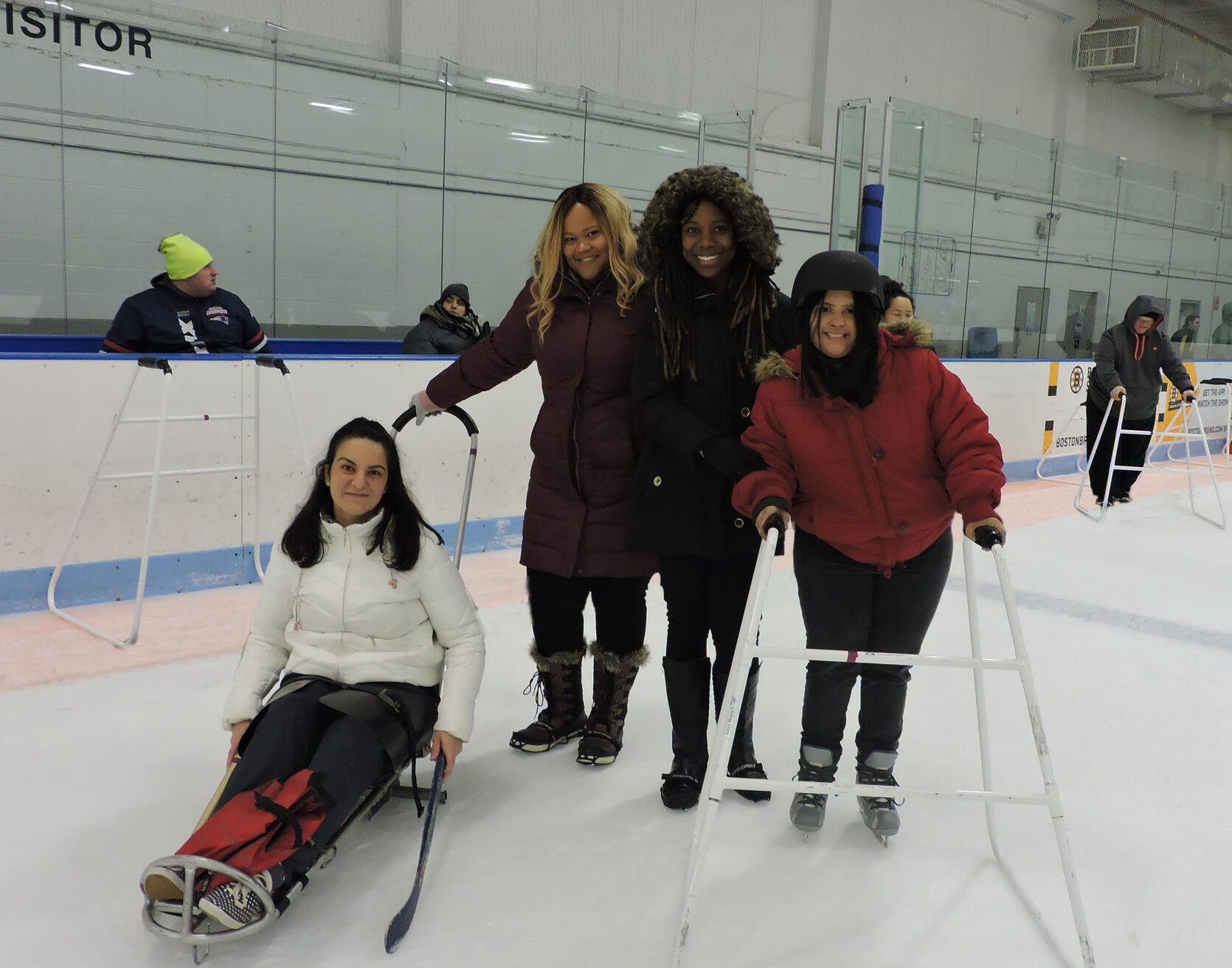 : Four women in winter coats are standing on the rink and smiling at the camera. One of the women is sitting in an ice sled with a handle on it. Two of the women are wearing ice grippers on their shoes. The fourth woman is using a pair of skates and an ice walker.